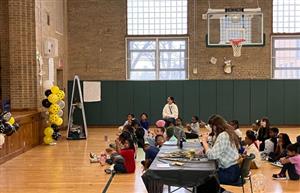 students and teachers sitting in a gym watching a spelling bee. 
